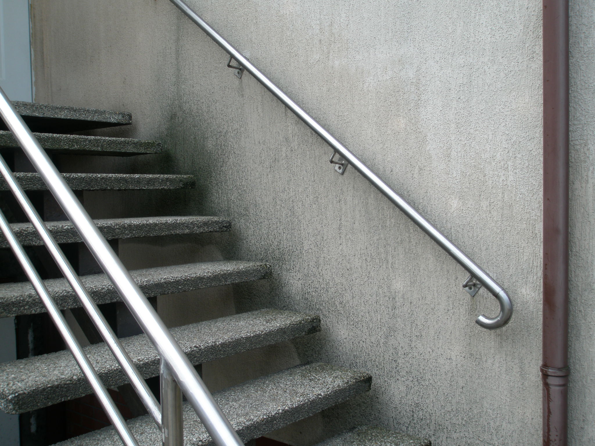 Betonowe schody zewnętrzne z metalową balustradą przymocowaną do szarej, chropowatej ściany budynku, obok rury spustowej.